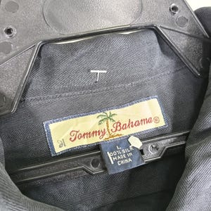Peut inclure: Gros plan sur une chemise en soie grise Tommy Bahama. La chemise est suspendue &agrave; un cintre noir. L'&eacute;tiquette Tommy Bahama est visible, ainsi qu'une &eacute;tiquette indiquant "100% Soie Fabriqu&eacute; en Chine."