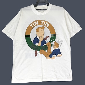 May include: A white short-sleeve t-shirt featuring a graphic of the character Tintin. The design includes a circular logo with the text TIN TIN SINCE 1929, showing a close-up of the character and a full-body illustration of Tintin falling down.