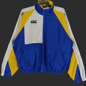 May include: A blue, white, and yellow zip-up jacket with the Canterbury logo on the chest. The jacket has a high collar and two front pockets. The sleeves and shoulders are white and yellow, while the body is blue.