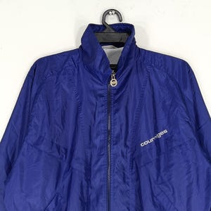 May include: A blue Courr&egrave;ges zip-up jacket with a high collar. The jacket has a white logo on the chest and a full-length zipper. The jacket is hanging on a black hanger.