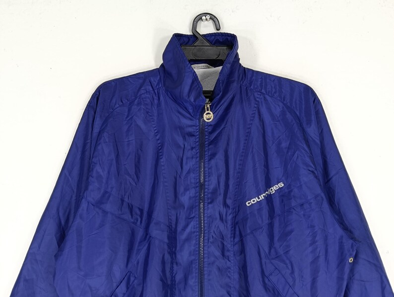 May include: A blue Courr&egrave;ges zip-up jacket with a high collar. The jacket has a white logo on the chest and a full-length zipper. The jacket is hanging on a black hanger.