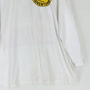 Puede incluir: Camiseta blanca de manga larga con un dise&ntilde;o circular amarillo y negro en el pecho izquierdo. El dise&ntilde;o dice "ADVENTURE". La camiseta est&aacute; hecha de un material suave.