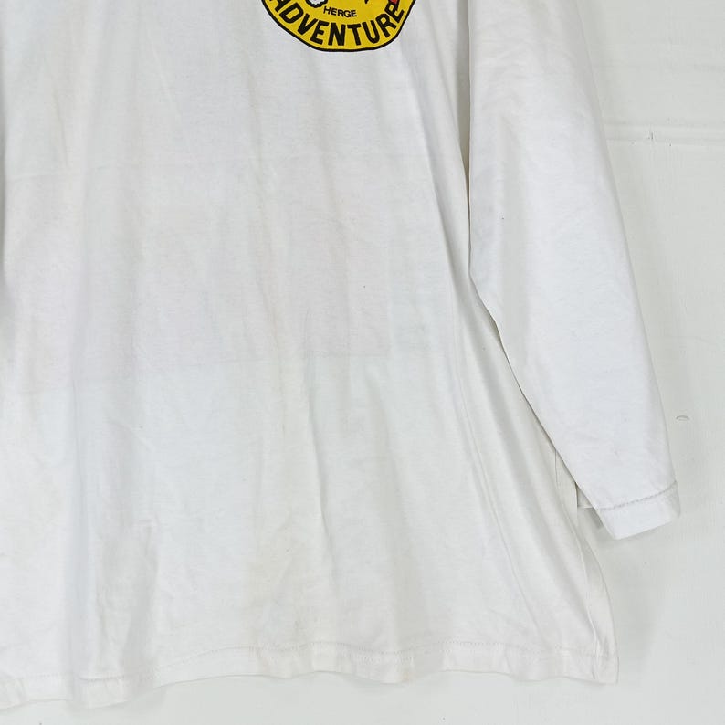Puede incluir: Camiseta blanca de manga larga con un dise&ntilde;o circular amarillo y negro en el pecho izquierdo. El dise&ntilde;o dice "ADVENTURE". La camiseta est&aacute; hecha de un material suave.
