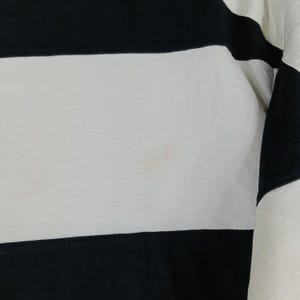 May include: Close-up of a black and white striped garment. The fabric has a slightly textured appearance. The stripes are horizontal, with alternating black and white bands. The garment appears to be a piece of clothing, possibly a shirt or top.