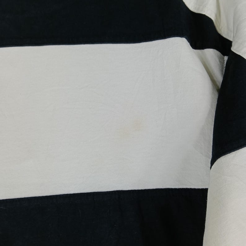 May include: Close-up of a black and white striped garment. The fabric has a slightly textured appearance. The stripes are horizontal, with alternating black and white bands. The garment appears to be a piece of clothing, possibly a shirt or top.