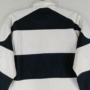 May include: A white and navy blue striped rugby shirt. The shirt has a white collar and white sleeves with navy blue accents. The back of the shirt is visible, showing the horizontal stripes and the collar.