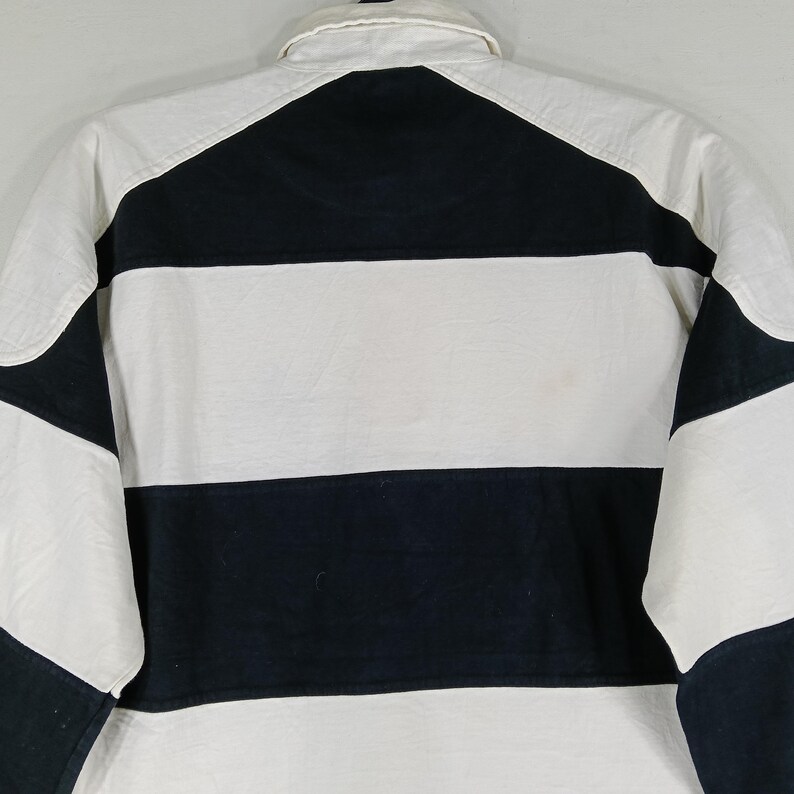 May include: A white and navy blue striped rugby shirt. The shirt has a white collar and white sleeves with navy blue accents. The back of the shirt is visible, showing the horizontal stripes and the collar.
