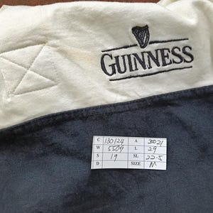 May include: Close-up of a cream and navy blue Guinness shirt. The Guinness logo, featuring a harp, is embroidered in black. The shirt has a stitched detail on the cream-colored shoulder. A size tag is visible.