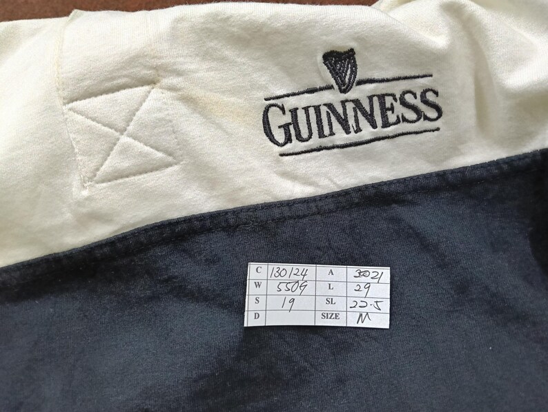 May include: Close-up of a cream and navy blue Guinness shirt. The Guinness logo, featuring a harp, is embroidered in black. The shirt has a stitched detail on the cream-colored shoulder. A size tag is visible.