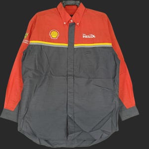 Puede incluir: Camisa abotonada de manga larga con diseño de bloques de color. La parte superior y las mangas son naranjas, mientras que el cuerpo es gris. La camisa presenta el logotipo de Shell, la palabra "HELIX" y otros gráficos.