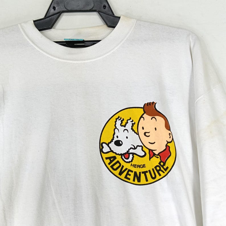 Puede incluir: Camiseta blanca con un gr&aacute;fico circular que presenta a un ni&ntilde;o y un perro de dibujos animados. El gr&aacute;fico es amarillo con texto negro "ADVENTURE" y "HERGE". El ni&ntilde;o tiene pelo casta&ntilde;o y una camisa roja.