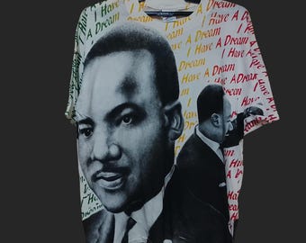 May include: White t-shirt featuring a black and white portrait of Martin Luther King Jr. with the words "I Have A Dream" in various colors. The shirt is hanging on a black hanger.