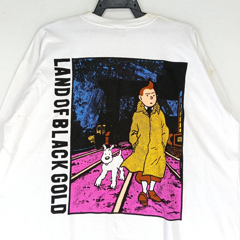 Puede incluir: Camiseta blanca de manga larga con un gr&aacute;fico del c&oacute;mic "Land of Black Gold". El dise&ntilde;o muestra un personaje con un abrigo amarillo con un perro blanco caminando por un camino rosa y morado.