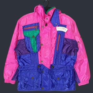 May include: A vibrant multi-colored jacket featuring a bright pink base with contrasting panels in royal blue, purple, and green. The jacket includes a high collar, multiple zippered pockets, a drawstring waist, and a white patch with the text WINDEX.