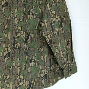 May include: A long-sleeved shirt with a camouflage pattern featuring green maple leaves and a brown bark-like background. The shirt has a buttoned cuff and a buttoned front.