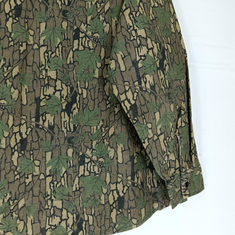 May include: A long-sleeved shirt with a camouflage pattern featuring green maple leaves and a brown bark-like background. The shirt has a buttoned cuff and a buttoned front.