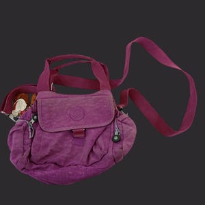 May include: A purple shoulder bag with a flap closure and a long, adjustable strap. The bag has multiple zippered compartments and a small, round logo on the front. The bag is made of a textured material and has a matching handle.