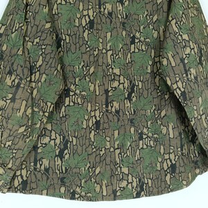 May include: A long-sleeved shirt with a camouflage pattern. The shirt features a bark-like design in shades of brown and tan, with green leaves scattered across the fabric. The overall effect is a natural, outdoorsy aesthetic.