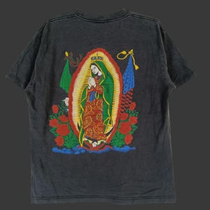 May include: Dark grey t-shirt with a vibrant graphic of the Virgin Mary. The design incorporates a halo, roses, and flags. The shirt has a vintage, washed-out look.