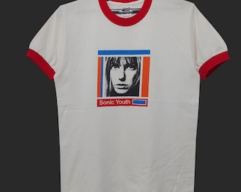 May include: White t-shirt with red trim on the collar and sleeves. The shirt features a black and white image of a woman's face within a blue and orange rectangle. The text "Sonic Youth" is below the image.