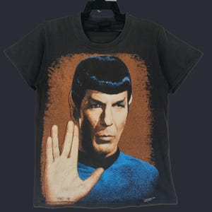 May include: A black short-sleeve t-shirt featuring a graphic print of Spock from Star Trek. The design shows Spock performing the Vulcan salute with a hand raised, set against a textured brown background, wearing a blue uniform shirt.