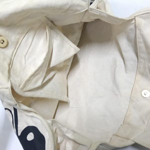 May include: An off-white canvas tote bag shown from an interior perspective, featuring a magnetic snap closure, a side pocket, and a partial black graphic print visible on the exterior lower corner of the fabric construction.