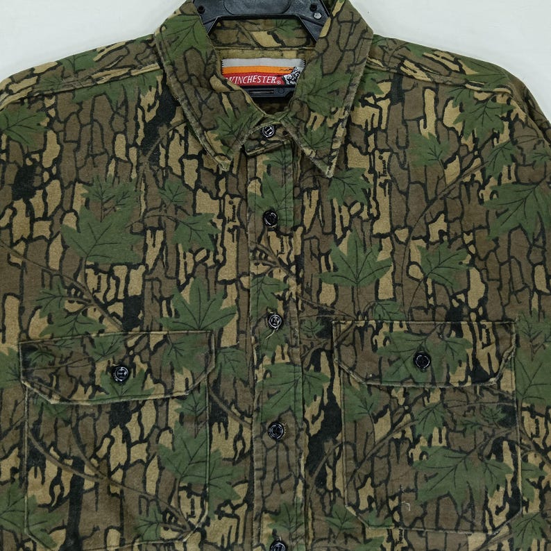 May include: A button-down shirt with a camouflage pattern in shades of green, brown, and black. The shirt features two chest pockets and a collar. The brand name "Winchester" is visible on the collar.