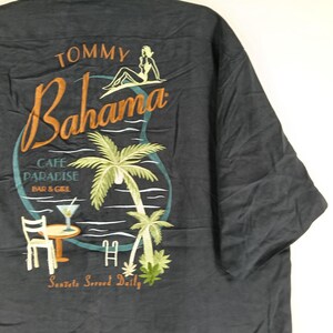 Peut inclure: Chemise boutonn&eacute;e gris fonc&eacute; avec un motif brod&eacute; color&eacute; au dos. Le motif comprend les mots "Tommy Bahama" en &eacute;criture orange, un palmier et le texte "Cafe Paradise Bar & Grill". La chemise porte &eacute;galement l'inscription "Sunsets Served Daily".