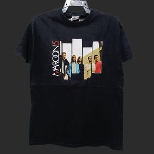 May include: Black t-shirt featuring the band Maroon 5. The shirt has a graphic of the band members with the band's name in white vertical text. The band members are standing in front of white vertical bars.