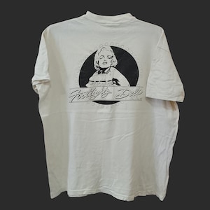 Puede incluir: Camiseta blanca con un gráfico en blanco y negro de una mujer en un círculo y el texto "Footlights Deli" debajo. El gráfico está en la parte posterior. La camiseta tiene mangas cortas y cuello redondo.