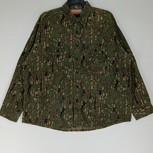 May include: A long-sleeved button-down shirt with a camouflage pattern featuring green leaves and brown bark-like textures. The shirt has two chest pockets and a collar. The overall color palette is earthy tones of green, brown, and black.