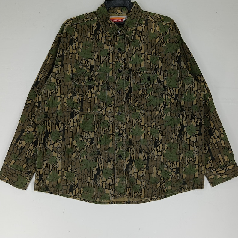 May include: A long-sleeved button-down shirt with a camouflage pattern featuring green leaves and brown bark-like textures. The shirt has two chest pockets and a collar. The overall color palette is earthy tones of green, brown, and black.