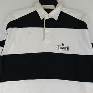 May include: A white and black rugby shirt with a collared neckline. The shirt features a horizontal black stripe across the chest and the Guinness logo embroidered on the front. The shirt has long sleeves and a button-down placket.