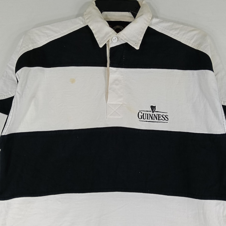 May include: A white and black rugby shirt with a collared neckline. The shirt features a horizontal black stripe across the chest and the Guinness logo embroidered on the front. The shirt has long sleeves and a button-down placket.