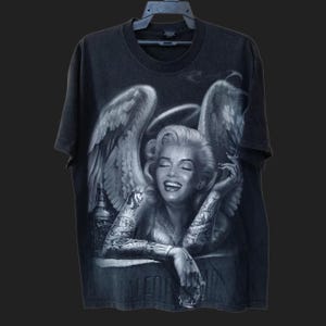 May include: Black t-shirt featuring a detailed graphic of a woman with angel wings, tattoos, and a halo. The image includes a bottle of whiskey. The design is in shades of gray and white.