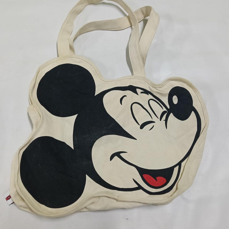 May include: A cream-colored canvas tote bag shaped like the head of Mickey Mouse. The bag features a large black graphic of Mickey Mouse's face with his signature ears, a smiling expression, and a red tongue, complete with two top handles.