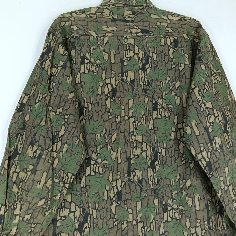May include: A long-sleeved button-down shirt with a camouflage pattern. The shirt features a brown and black bark-like design with green maple leaves. The shirt is made of a durable fabric and has a classic collar.