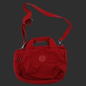 May include: A red nylon crossbody bag with a top handle and adjustable shoulder strap. The bag features a small, round logo patch on the front and silver-tone hardware. The bag is designed for everyday use.