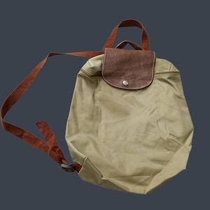 May include: A beige nylon backpack featuring a brown textured leather flap with a gold-tone snap button closure. The bag includes brown fabric shoulder straps and a top handle, with a zip closure along the top edge of the main compartment.