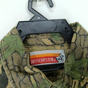 May include: A camouflage-patterned shirt with green, brown, and tan hues, hanging on a black hanger. The shirt features a "Winchester" label with a logo of a person riding a horse.