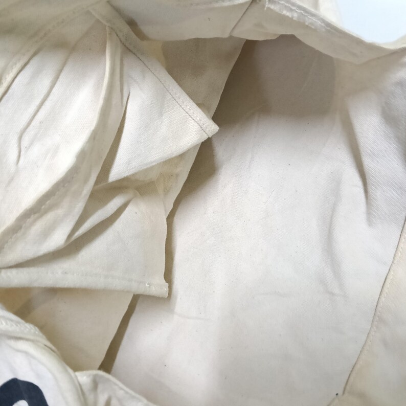 May include: The interior of an off-white canvas tote bag featuring a single open side pocket. The fabric shows a natural woven texture with visible stitching along the seams and the pocket edge. The bag is empty and open, revealing the light-colored lining.