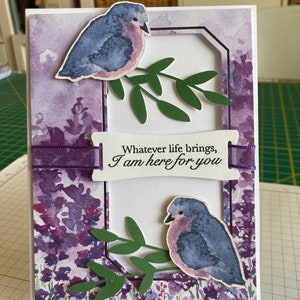 May include: A purple and white card with a watercolor background featuring two birds and green leaves. The card has a white banner with the text "Whatever life brings, I am here for you".