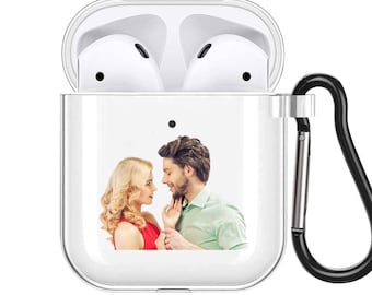 Puede incluir: Funda transparente para AirPod con mosquetón negro. La funda presenta una imagen impresa de una pareja. La funda está abierta, revelando los Airpods blancos en el interior. Una segunda imagen de la pareja está en la esquina superior derecha.