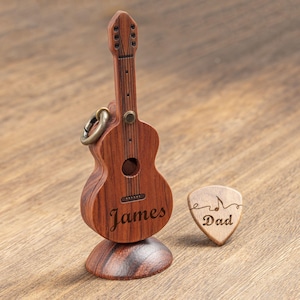 Puede incluir: Una guitarra de madera en miniatura con el nombre "James" grabado en el cuerpo, acompañada de una púa con la inscripción "Dad". La guitarra tiene un anillo de metal y se apoya en una base de madera.