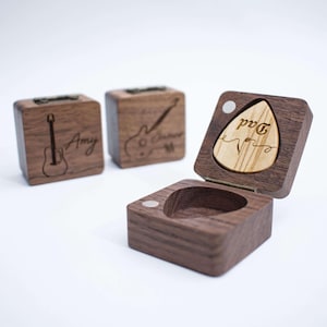 Könnte beinhalten: Gitarrenplektrum-Box aus Holz mit einem Plektrum mit der Aufschrift "Dad". Zwei zusätzliche Holzkisten mit Gitarrenillustrationen und Namen sind ebenfalls sichtbar. Die Boxen sind aus dunklem Holz und haben einen Klappdeckel.