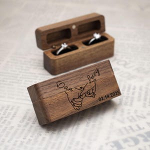 May include: A wooden ring box with two compartments for rings. The box is engraved with the names "Dan & Lucy" and the date "02.14.2025". The lid of the box features a design of two hands holding each other.