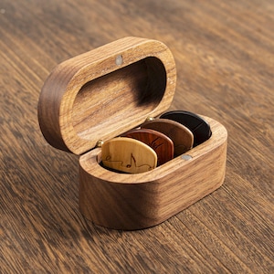 May include: An oval wooden box with a hinged lid containing four guitar picks. The picks are made of different shades of wood, ranging from light tan to dark brown and black. The front pick features a small engraved musical note design.