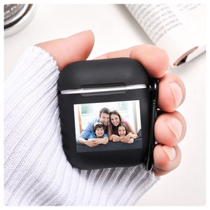 K&ouml;nnte beinhalten: Schwarzes AirPods-Etui mit einem Familienfoto. Das Etui wird in einer Hand mit einem wei&szlig;en Pullover gehalten. Das Foto zeigt eine l&auml;chelnde vierk&ouml;pfige Familie. Das Etui hat einen schwarzen Karabinerhaken.