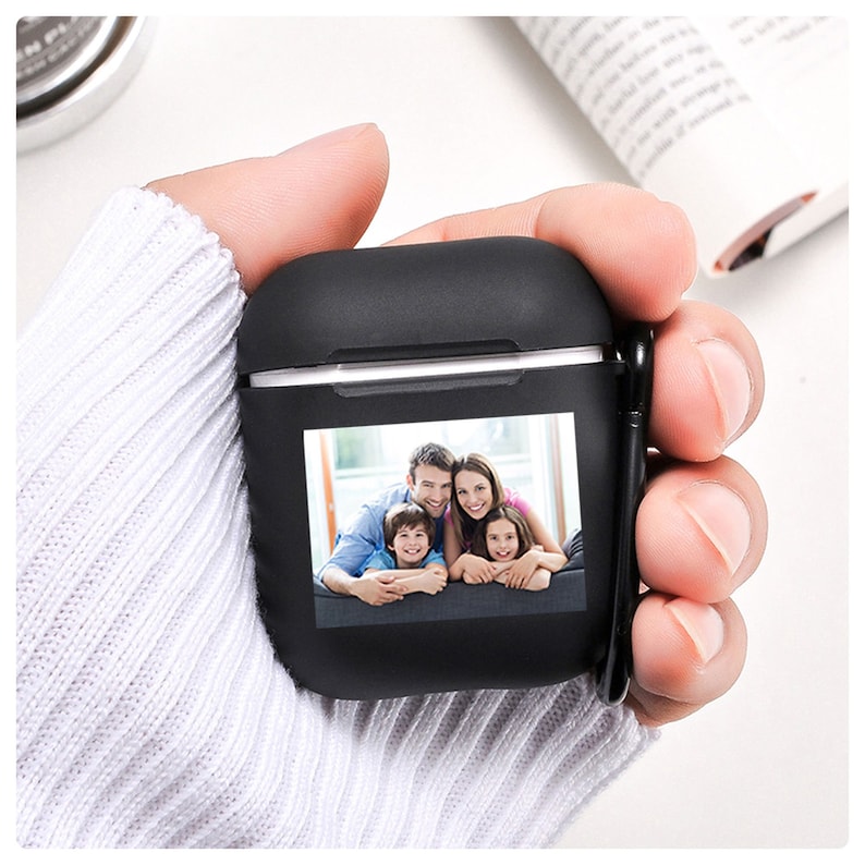 K&ouml;nnte beinhalten: Schwarzes AirPods-Etui mit einem Familienfoto. Das Etui wird in einer Hand mit einem wei&szlig;en Pullover gehalten. Das Foto zeigt eine l&auml;chelnde vierk&ouml;pfige Familie. Das Etui hat einen schwarzen Karabinerhaken.