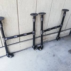 May include: Three black metal pipe table legs with wheels. Each leg has a round metal plate at the top and a black pipe extending down to a wheel.